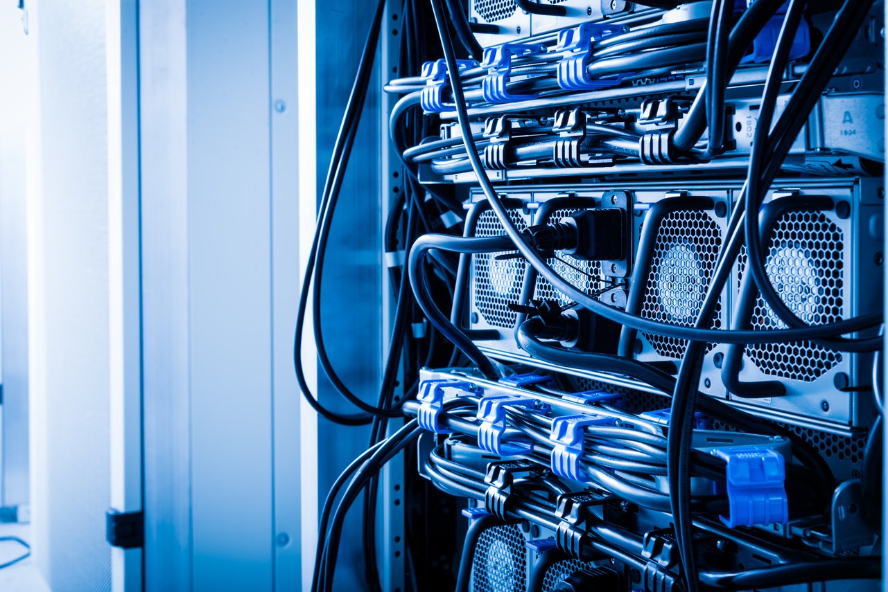 A imagem mostra um ambiente tecnológico, provavelmente o interior de um data center ou sala de servidores. Nela, é possível ver racks metálicos com vários equipamentos de TI conectados por cabos de energia e de rede organizados com presilhas azuis. A iluminação em tons de azul reforça a atmosfera de alta tecnologia e resfriamento, transmitindo uma sensação de ambiente controlado e de infraestrutura crítica. Essa cena representa o núcleo físico da computação e da conectividade digital, onde dados e aplicações corporativas são processados e armazenados.