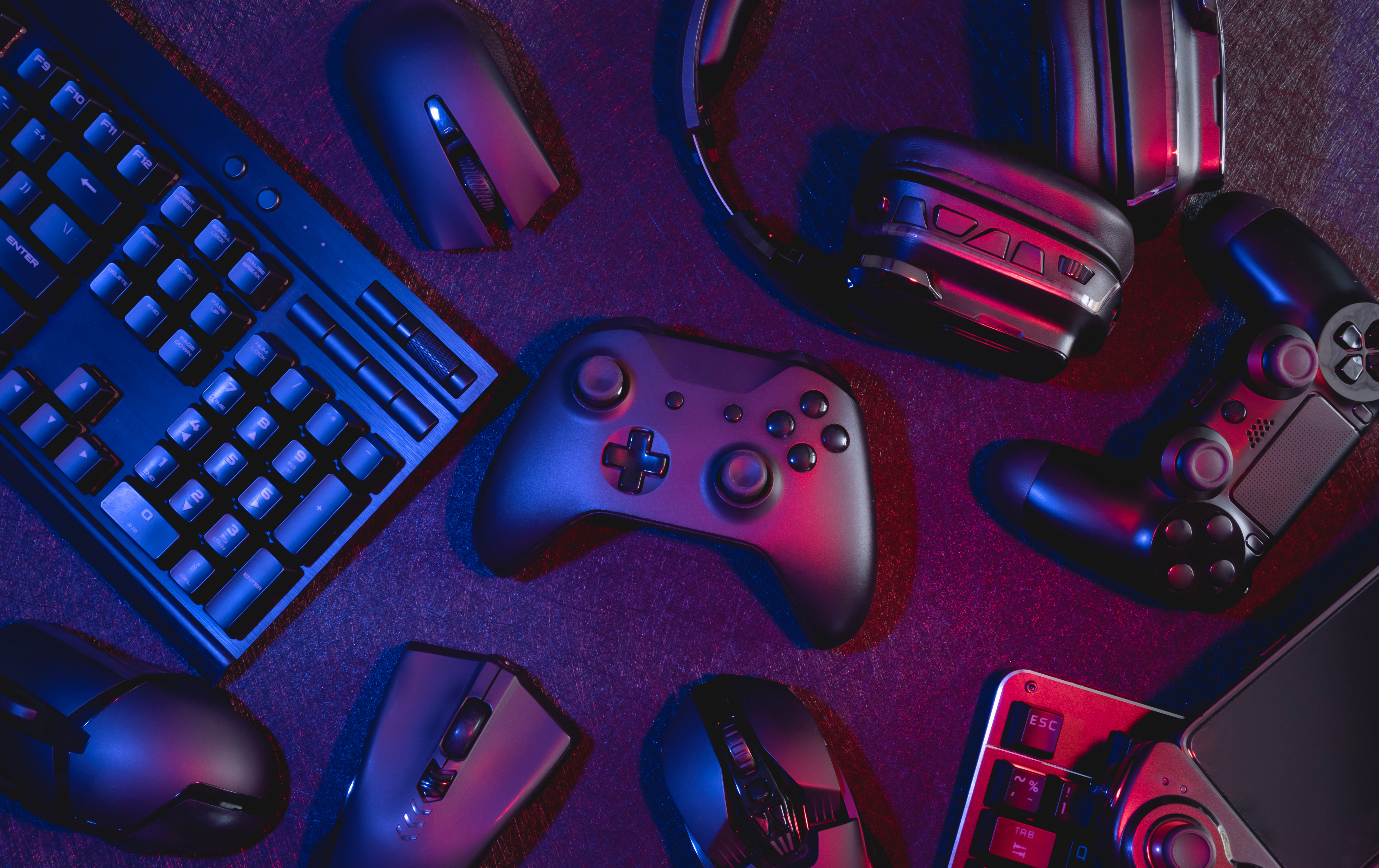 Vista superior de uma mesa gamer com teclado mecânico, mouses, headsets e controles de videogame iluminados por luzes em tons de azul e vermelho. A imagem representa setup gamer, acessórios para jogos, periféricos de alta performance, eSports, PC gaming e consoles, com foco em tecnologia, entretenimento digital e experiência imersiva para jogadores.