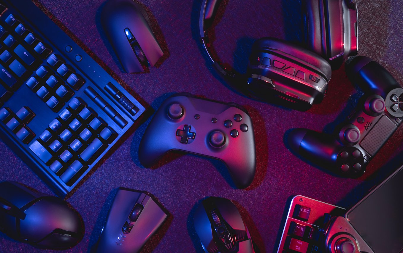 Vista superior de uma mesa gamer com teclado mecânico, mouses, headsets e controles de videogame iluminados por luzes em tons de azul e vermelho. A imagem representa setup gamer, acessórios para jogos, periféricos de alta performance, eSports, PC gaming e consoles, com foco em tecnologia, entretenimento digital e experiência imersiva para jogadores.