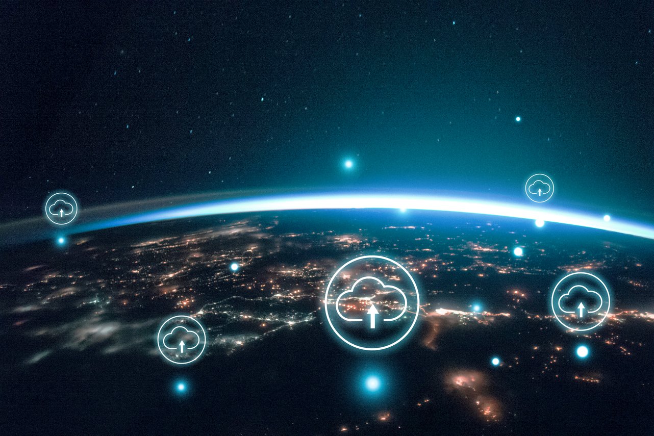 A imagem mostra a curvatura da Terra vista do espaço, como se a cena tivesse sido capturada a partir de um satélite em órbita. O planeta aparece parcialmente iluminado, com o horizonte azul brilhante destacando a atmosfera e inúmeras luzes espalhadas pela superfície, sugerindo cidades à noite. O fundo é escuro, repleto de estrelas, reforçando a sensação de profundidade e escala espacial. Sobre a paisagem terrestre, há ícones gráficos de nuvem com setas apontando para cima, distribuídos em diferentes pontos do globo. Esses símbolos representam upload, armazenamento ou conexão em nuvem, sugerindo infraestrutura de cloud computing global, troca de dados e conectividade digital entre regiões do mundo. A composição combina elementos reais (a Terra, inspirada em imagens da NASA) com gráficos tecnológicos, transmitindo a ideia de rede, tecnologia, dados e transformação digital em escala planetária.